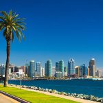 Downtown San Diego skyline view with the San Diego Bay, a waterfront walkway, and a palm tree in the foreground, and a nearly clear deep blue sky in the background.